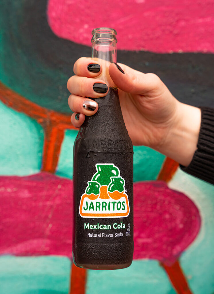 A close-up of a hand holding a chilled Jarritos Mexican Cola bottle in front of a vibrant, colorful mural. The bold label and condensation on the glass emphasize the soda’s refreshing taste and lively personality, perfect for enjoying authentic Mexican flavor in any setting.