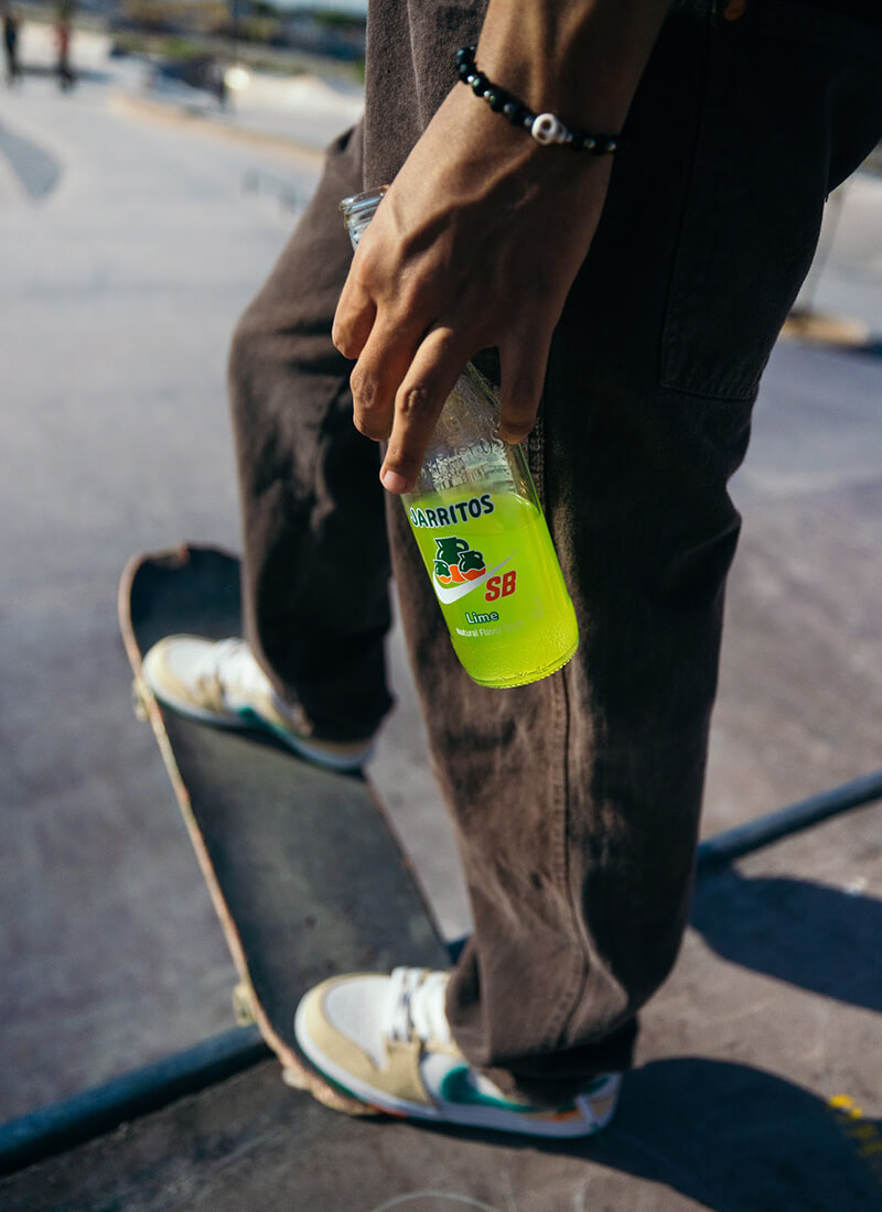 Close-up of a skateboarder holding a limited-edition Jarritos SB Lime soda bottle, wearing Nike SB x Jarritos sneakers—capturing the bold energy of this collaboration through urban streetwear, vibrant flavor, and skate culture style.