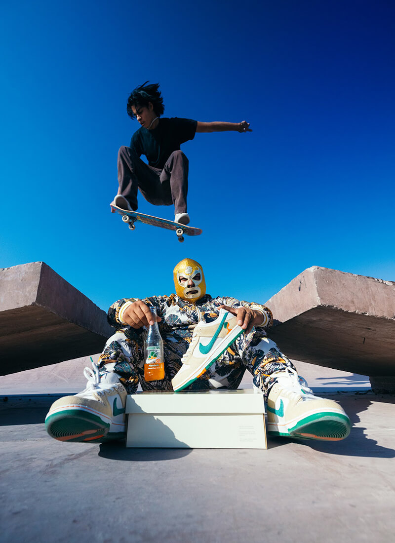 A masked skater sits with a Jarritos soda and Nike SB Dunk Low sneaker from the Jarritos x Nike collaboration, while another skater performs a trick in midair—blending street style, action, and bold Mexican-inspired design in an iconic urban scene.