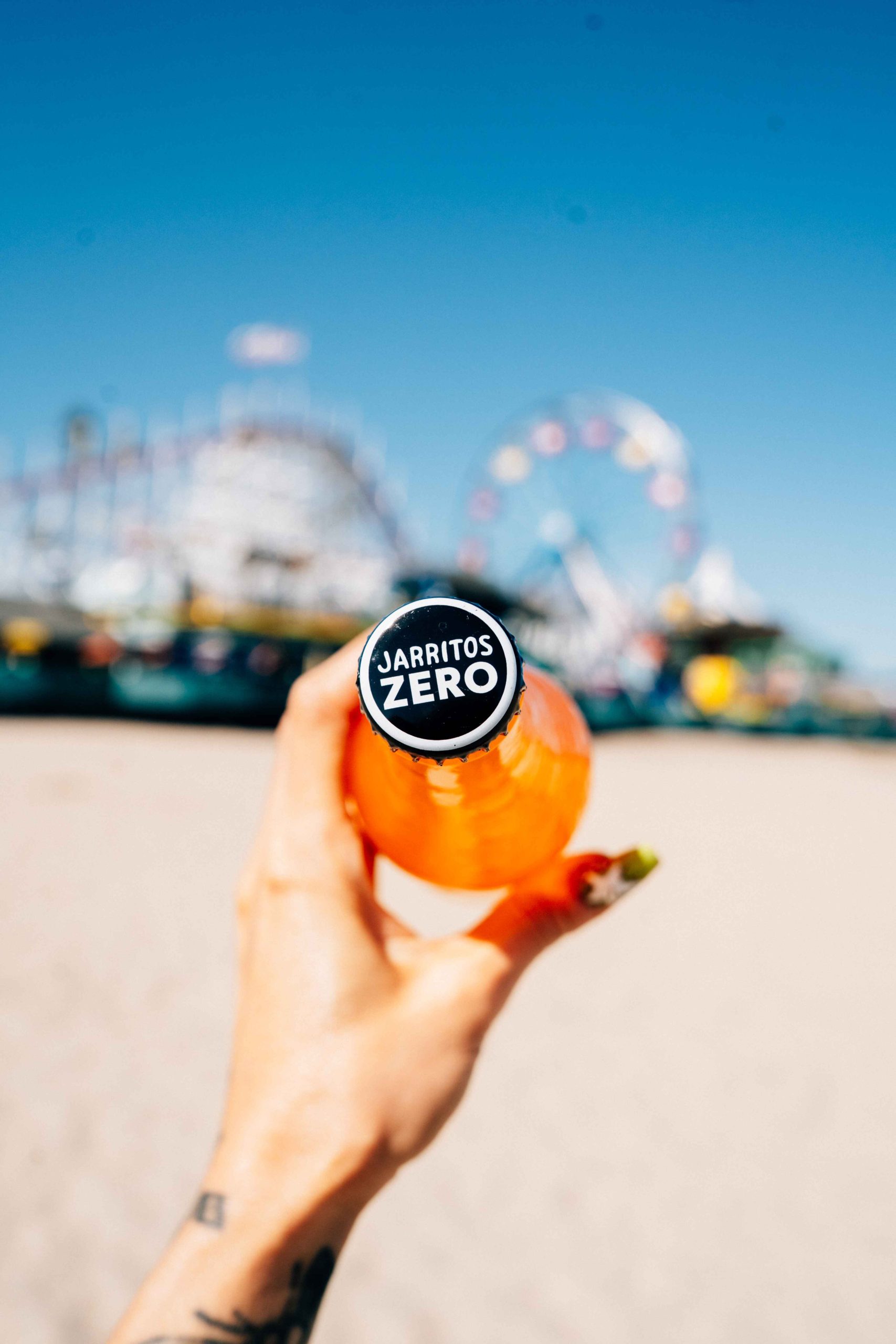 Close-up of a hand holding a cold Jarritos Zero soda bottle at the beach, with the cap in focus and a colorful amusement park blurred in the background—capturing a vibrant, carefree summer vibe with a refreshing, zero-calorie drink.