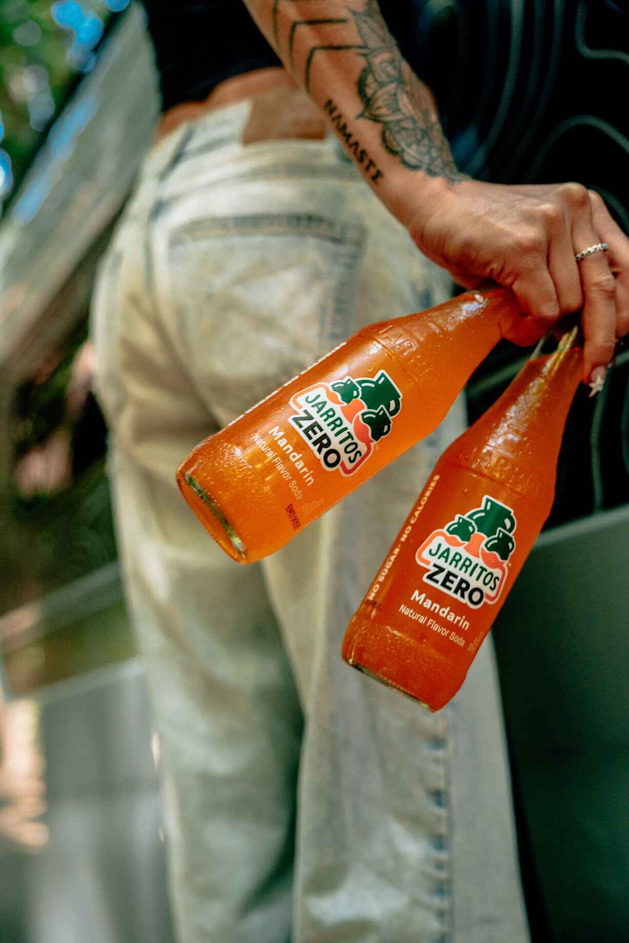 A person holds two chilled bottles of Jarritos Zero Mandarin soda, showcasing the vibrant orange drink and bold branding—highlighting its zero sugar, zero calorie formula in a refreshing, stylish street setting.