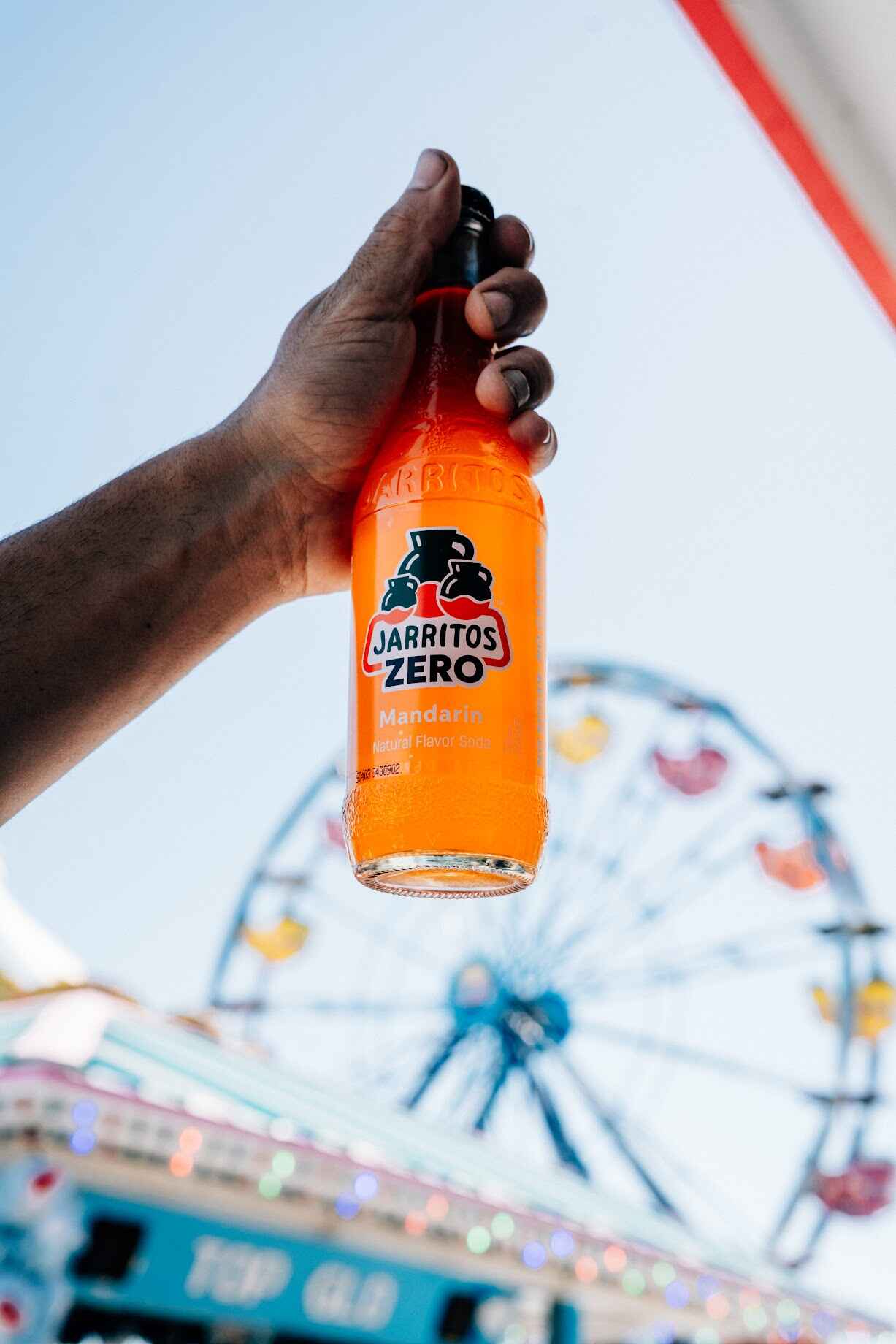 A hand holds up a bottle of Jarritos Zero Mandarin soda against a bright sky, with a colorful Ferris wheel and carnival lights in the background—capturing the fun, flavor, and refreshment of a zero-calorie drink on a sunny day.