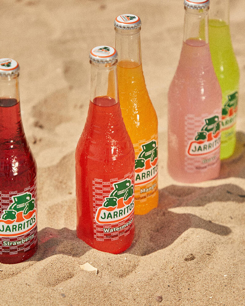 A colorful lineup of Jarritos sodas featuring Strawberry, Watermelon, Mango, and Lime flavors is displayed on the sand, showcasing the exclusive collaboration with Kith Treats. The limited-edition labels with “KITH” branding highlight a refreshing summer vibe and bold brand partnership.