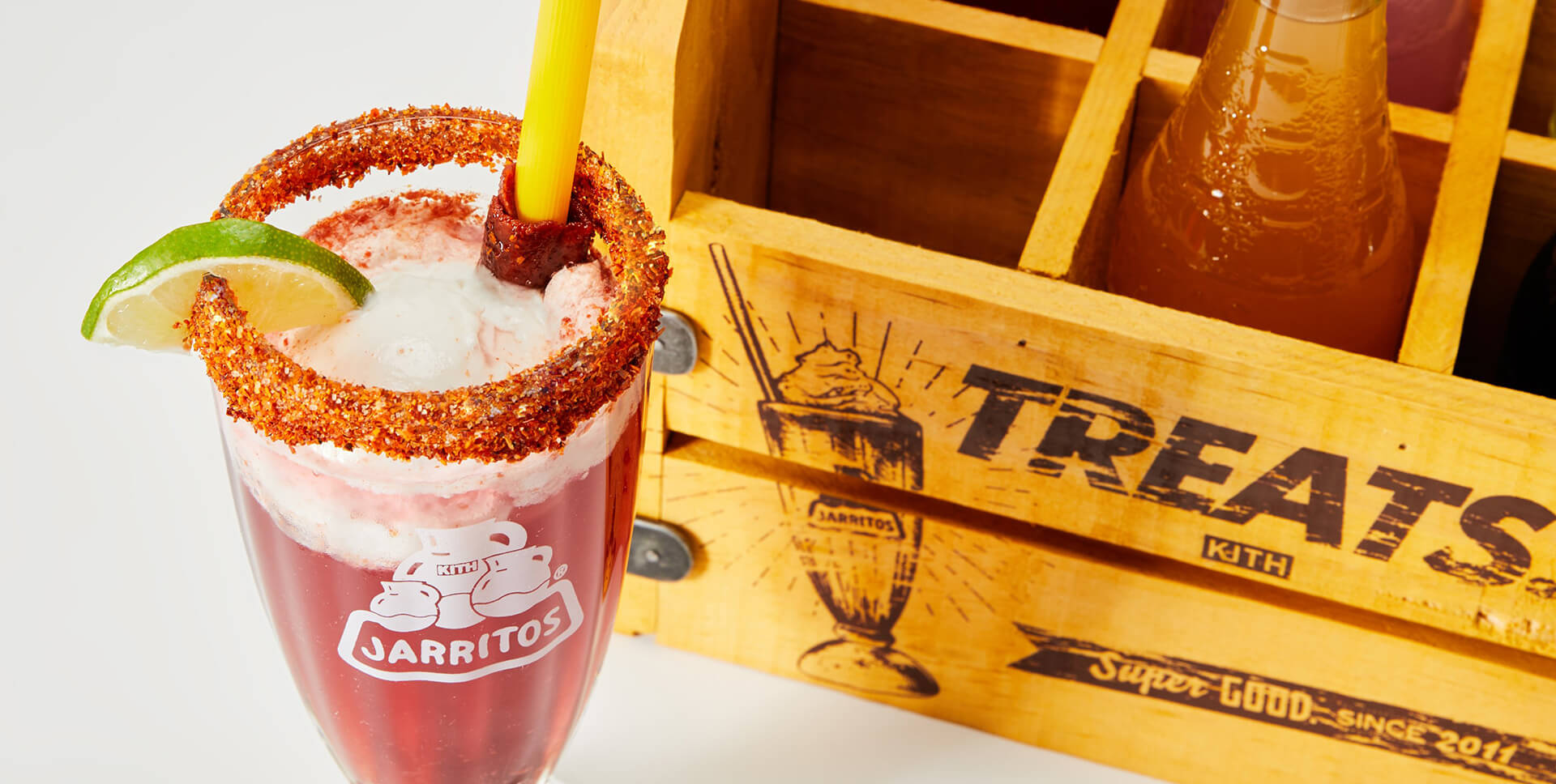 A vibrant Jarritos float topped with vanilla ice cream, chamoy rim, lime wedge, and tamarind candy is served in a branded glass. Next to it sits a wooden crate labeled “Kith Treats,” highlighting the exclusive collaboration between Kith Treats and Jarritos.