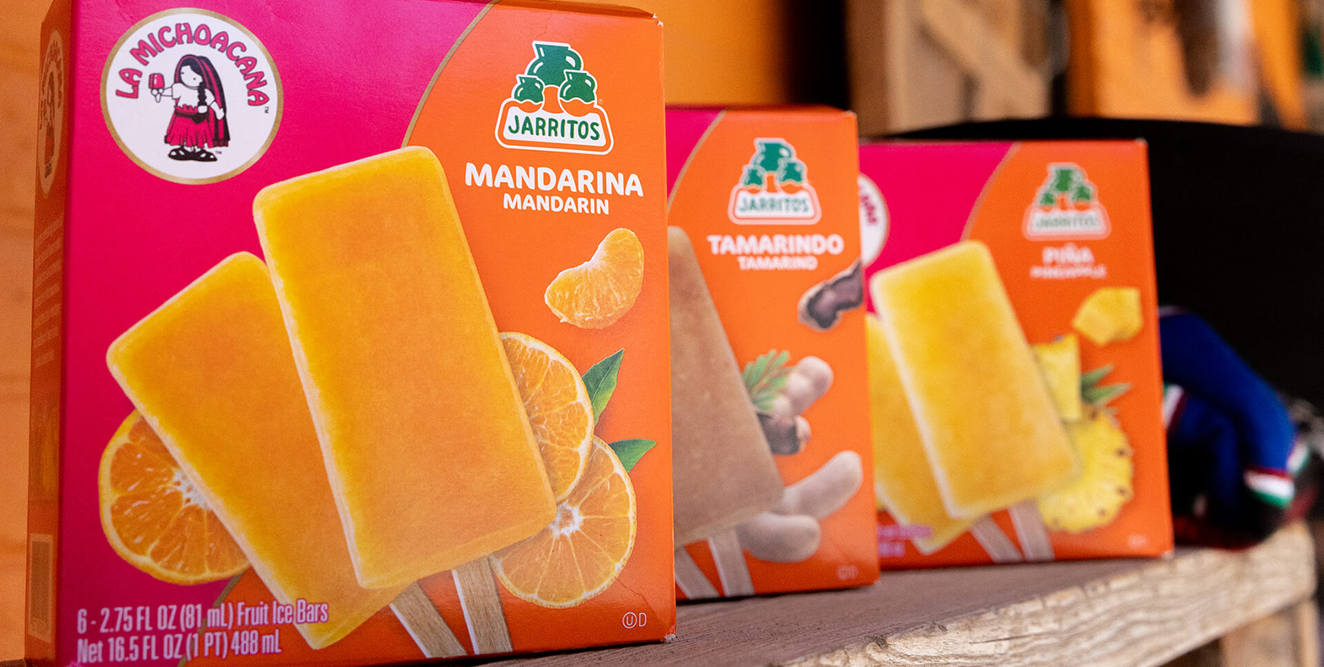 Close-up of La Michoacana and Jarritos collaborative popsicle boxes featuring Mandarina, Tamarindo, and Piña flavors—brightly packaged and displayed on a rustic wooden shelf, blending two iconic Mexican brands in a flavorful frozen treat.
