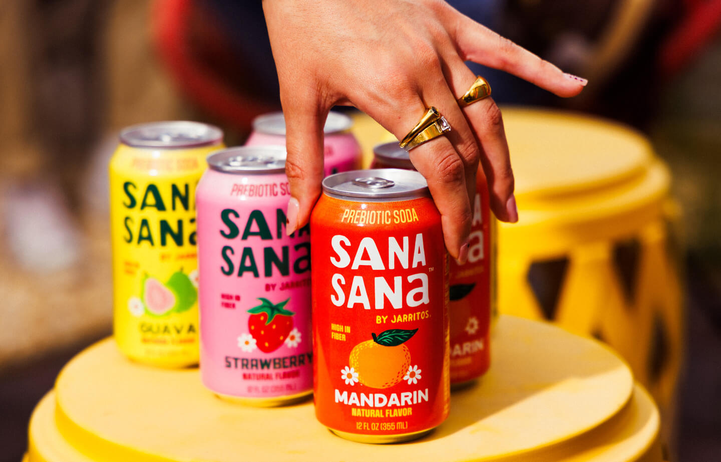 Hand reaching for a Sana Sana by Jarritos mandarin prebiotic soda can, surrounded by guava and strawberry flavors, highlighting the vibrant and better-for-you Jarritos Familia line.