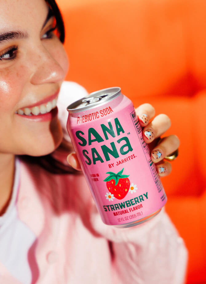 Smiling woman holding a Sana Sana by Jarritos strawberry prebiotic soda can, showcasing the refreshing, fruit-forward flavor and vibrant Jarritos Familia collection.