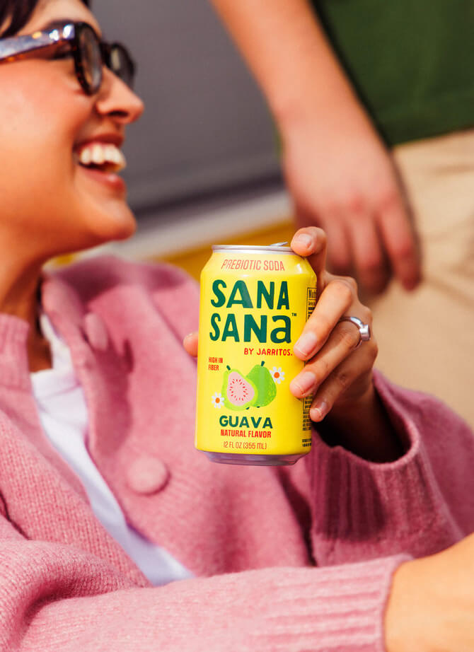 Woman wearing sunglasses holding a Sana Sana by Jarritos guava prebiotic soda can, highlighting the refreshing tropical flavor and colorful Jarritos Familia lineup.